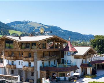 A charming hotel in alpine style with wooden cladding. The surroundings offer an impressive mountain backdrop and a green garden.
