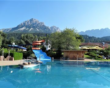 A beautiful pool with a water slide and a view of the mountains. Surrounded by green trees and sunny weather.