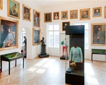 A museum with historical portraits on the walls. In the center stands a display case with a uniform and a helmet.
