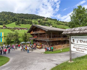 Ein traditionelles Holzhaus in der Berglandschaft mit vielen Besuchern. Im Vordergrund steht ein Schild für den Handwerksmarkt.