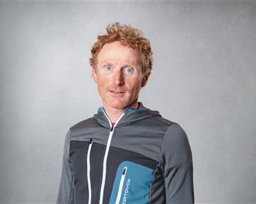 A man with red hair is wearing a sporty jacket. He stands in front of a neutral background and looks directly at the camera.