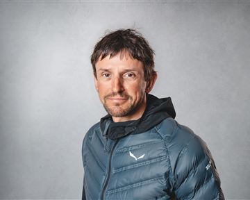 A man with short hair is wearing a blue down jacket. He is standing in front of a neutral, gray background.