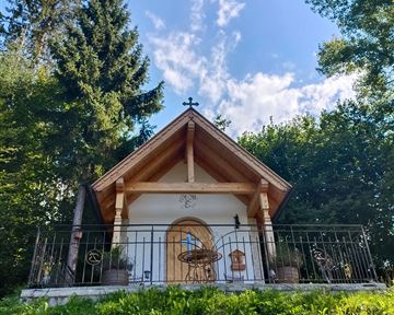 A small chapel in a green meadow, surrounded by trees. The sky is blue with some clouds.