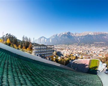 Ein Blick auf die Stadt mit Bergen im Hintergrund und klarem blauen Himmel. Im Vordergrund sind die Schanzenanlagen und die umliegende Landschaft zu sehen.