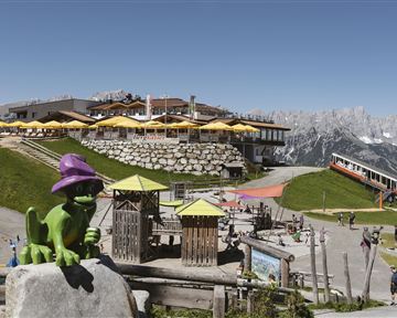 Eine Berglandschaft mit einem Restaurant und sonnigen Terrassen. Im Vordergrund steht eine grüne Figur mit einem lila Hut und Spielgeräte für Kinder.