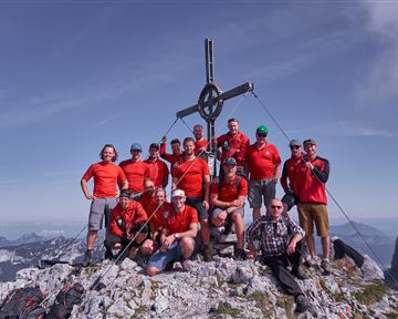Eine Gruppe von zehn Personen steht auf einem Berggipfel, umgeben von Felsen und einem klaren Himmel. Sie tragen rote T-Shirts und posieren fröhlich vor einem Kreuz.