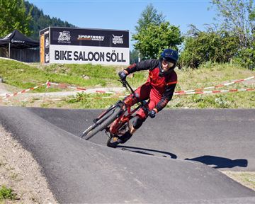 A mountain biker is riding on a winding track. In the background, the sign "Bike Saloon Söll" is visible.