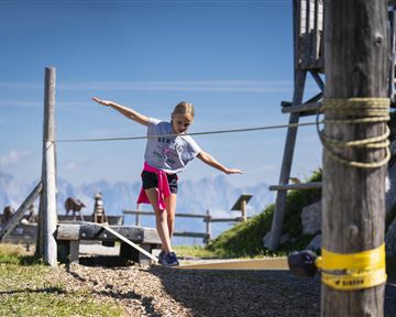 A girl is balancing on a rope in the mountains. In the background, a beautiful landscape and a wooden structure are visible.