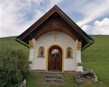 A small, charming chapel with a wooden roof and colorful windows stands on a green meadow. The sky is slightly cloudy.