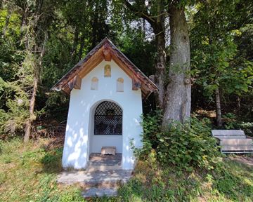 Eine kleine Kapelle steht in einer bewaldeten Umgebung. Neben der Kapelle befindet sich eine Holzbank.