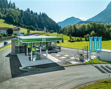 Eine moderne Tankstelle in einer malerischen Berglandschaft. Die Umgebung ist grün mit Hügeln und blauen Bergen im Hintergrund.