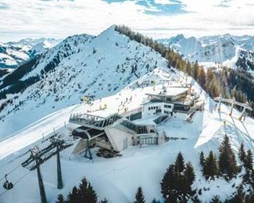 Eine schneebedeckte Berglandschaft mit Skiliften und einer modernen Bergstation. Im Hintergrund sind majestätische Berggipfel und ein klarer Himmel zu sehen.