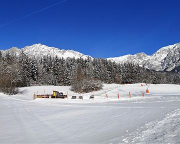 A snowy landscape with high mountains in the background. Some shoveling and seating areas are visible, surrounded by snow-covered trees.