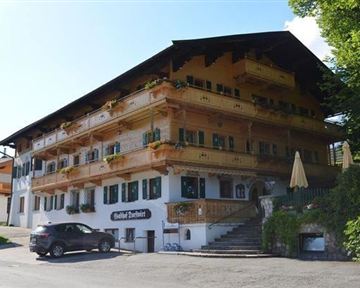 Ein charmantes Gebäude im bayerischen Stil mit großen Fenstern und Balkonen. Vor dem Haus stehen Autos und Sonnenschirme auf der Terrasse.