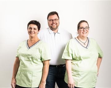 Ein sympathisches Team von drei Personen, zwei in grünen Arbeitskleidung und eine Person in einem weißen Hemd. Sie stehen vor einem neutralen, hellen Hintergrund.