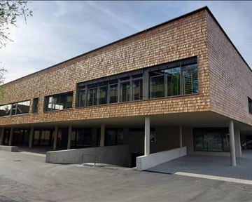 Ein modernes Gebäude mit Holzverkleidung und großen Fenstern. Es liegt in einer grünen Umgebung und hat eine aufgeräumte Architektur.