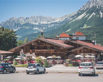 A traditional chalet in the mountains with blooming flowers and outdoor seating. In the background, majestic mountains and a clear blue sky are visible.