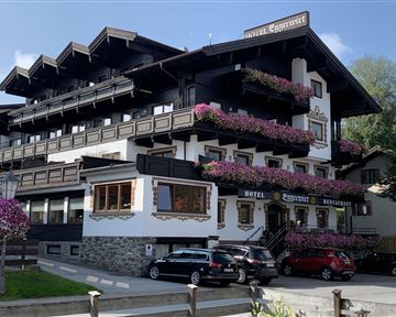 A charming hotel with traditional architecture and colorful flower boxes. The outdoor area is well-maintained and several cars are parked in front of it.