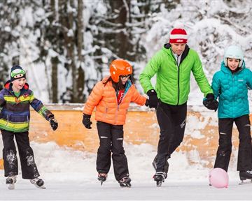 A cheerful group of children and one adult ice skating on an ice rink in the snow. They are wearing colorful winter clothing and having a lot of fun gliding together.