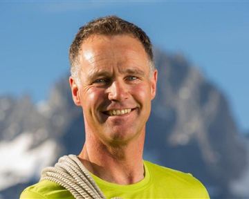 A smiling man is wearing a rope over his shoulder. In the background, mountains and a clear sky are visible.