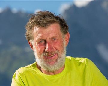 A smiling man with gray hair and a beard is wearing a bright yellow T-shirt. Beautiful mountains are visible in the background.