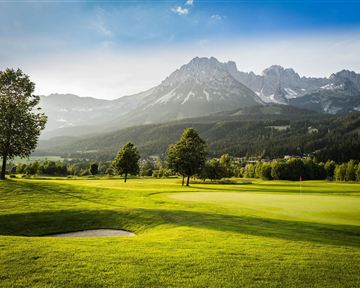Eine malerische Golfanlage mit sattem Grün und sanften Hügeln. Im Hintergrund erheben sich majestätische Berge unter einem klaren Himmel.