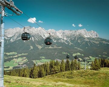 Eine Seilbahn fährt über eine grüne Landschaft mit Bergen im Hintergrund. Der Himmel ist klar und blau.