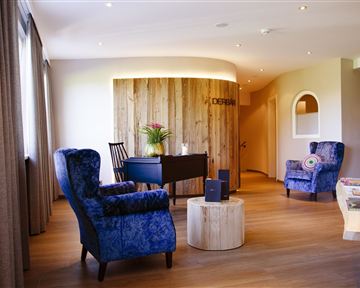 An elegant reception area with blue armchairs and a round wooden table. In the background, there is a wood paneling with plants.