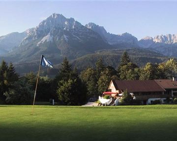 Ein schöner Golfplatz mit einem Flaggenstock im Vordergrund. Im Hintergrund sind majestätische Berge und ein typisches alpenländisches Haus zu sehen.