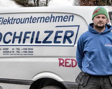 Ein Handwerker steht vor einem Lieferwagen mit der Aufschrift "Elektrounternehmen Hochfilzer". Der Mann trägt einen blauen Hoodie und eine grüne Mütze.