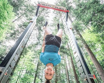 A young man is hanging upside down on a training device in the woods. The surroundings are green and filled with light.