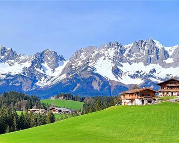 A picturesque mountain landscape with snow-capped peaks and lush green meadows. In the foreground, traditional wooden houses and a path can be seen.
