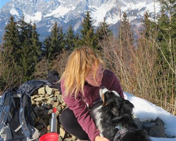 Ein Mann sitzt auf einem Weg und streichelt seinen Hund. Im Hintergrund sind hohe Berge und ein grünes Tal zu sehen.