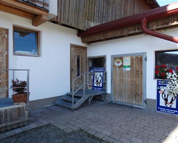 Ein Gebäude mit Holzverkleidung und zwei Türen. Vor der Eingangstreppe stehen Pflanzen und Plakate.