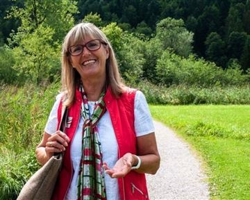 A friendly woman smiles on a hiking path surrounded by green nature. In the background, trees and a sunny landscape can be seen.