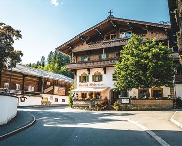 Ein traditionelles Gebäude im alpinen Stil mit holzverkleideter Fassade. Umgeben von Bäumen und einem klaren blauen Himmel.
