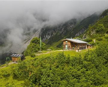 Eine rustikale Hütte umgeben von grünen Wiesen und Bergen. Der Himmel ist bewölkt und die Landschaft wirkt ruhig und idyllisch.