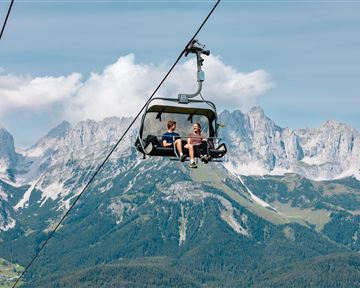 Two people are sitting in a gondola on their way over the mountains. The landscape consists of green forests and majestic peaks under a clear sky.