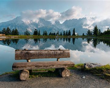 A wooden bench stands by the shore of a calm lake, surrounded by mountains. The sky is partly cloudy and reflects in the water.