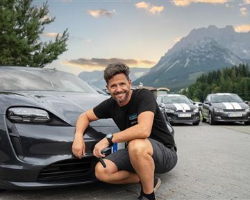 Ein Mann posiert lächelnd neben einem modernen Auto in einer bergigen Landschaft. Im Hintergrund sind weitere Autos und ein schöner Himmel zu sehen.