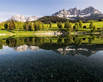 A picturesque landscape with a tranquil lake and gentle hills. In the background, majestic mountains rise under a clear sky.