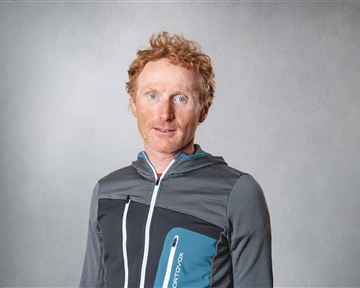 Ein Mann mit roten Haaren trägt eine sportliche Jacke. Er steht vor einem neutralen Hintergrund und schaut direkt in die Kamera.