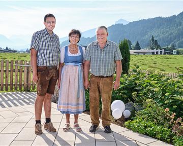 Eine Gruppe von sechs Personen in traditionellen Trachten steht vor einer beeindruckenden Berglandschaft. Im Hintergrund sind grüne Wälder und hohe Berge sichtbar.