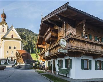 A traditional old inn with wooden decoration and green shutters stands next to a beautiful church. In the background, green hills and a clear sky are visible.