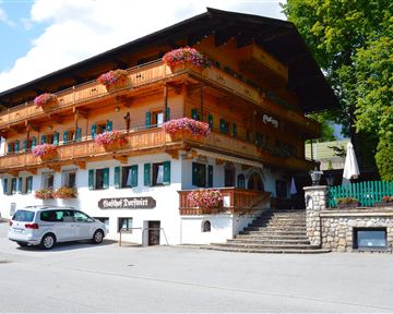 A traditional wooden house with balconies decorated with flowers. Additionally, a green surrounding and a parking lot in front.