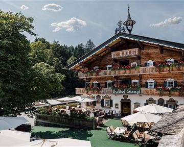 Ein traditionelles Alpenhaus mit blühenden Balkonen und einem roten Teppich vor dem Eingang. Umgeben von grünen Bäumen und einer einladenden Terrasse.