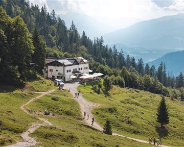 Ein malerisches Berghaus umgeben von grünen Wiesen und Bäumen. Der Weg führt zu dem Gebäude, das einen atemberaubenden Ausblick auf die Landschaft bietet.