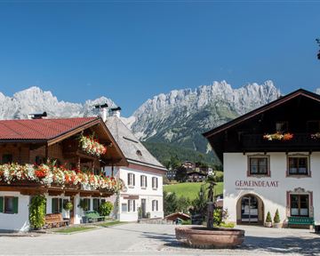 A picturesque alpine landscape with traditional wooden houses. In the background, majestic mountains rise under a clear blue sky.