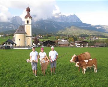 A picturesque rural scene with three people in traditional clothing standing in front of a church. In the foreground, two cows can be seen on a green meadow, surrounded by mountains.