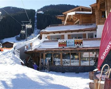 Ein Schneegebiet mit einem Bergrestaurant und Skiausleihe. Im Hintergrund sind Skilifte und schneebedeckte Berge zu sehen.